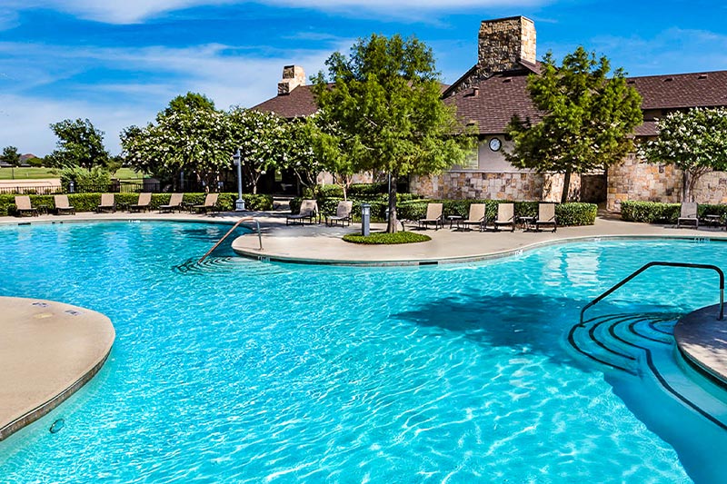 View of the outdoor pool and patio at Robson Ranch - Texas with landscaping and a clubhouse in the background.