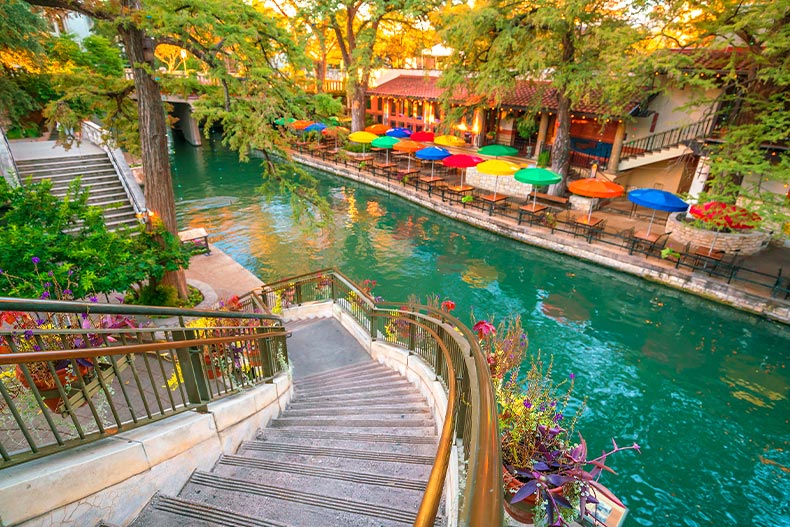 A staircase, trees, and a cafe with colorful umbrellas over outdoor seating at the San Antonio River Walk in Texas