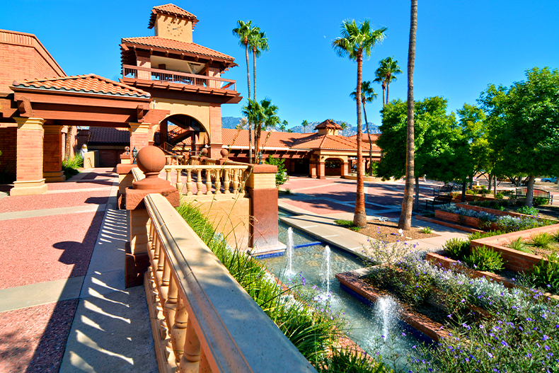 A railing leading up to the orange and tan exterior of the amenity center in Sun City Oro Valley with a fountain, trees, and flowers all around
