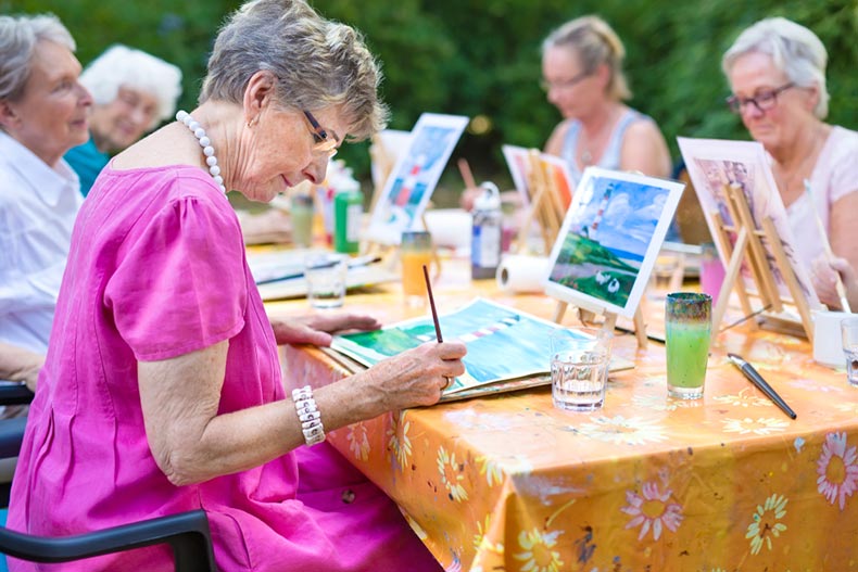 Stylish senior lady painting in art class with friends outdoors at a table in the garden