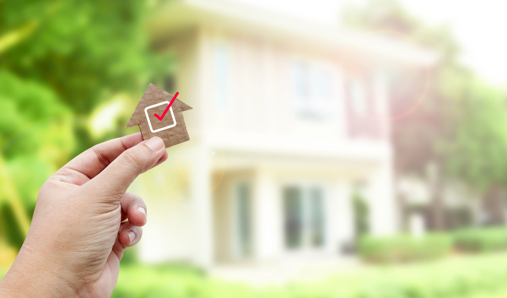 A hand holding a brown paper cutout of a house with a checkmark to symbolize a home inspection