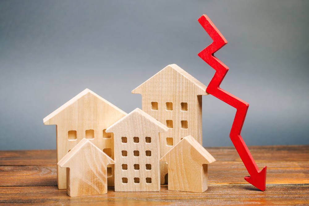 Five miniature wooden houses sitting on a wooden table beside a red arrow pointing down to symbolize lower mortgage interest rates