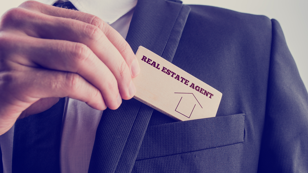 A real estate agent in a black suit putting a small wooden piece with real estate agent text into his front pocket