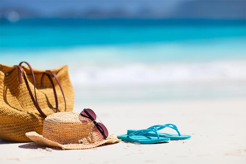 A straw hat, a bag, sun glasses, and flip flops on a tropical beach