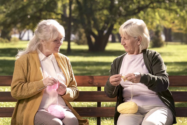 Two older women sitting on a park bench in the summertime and crocheting together