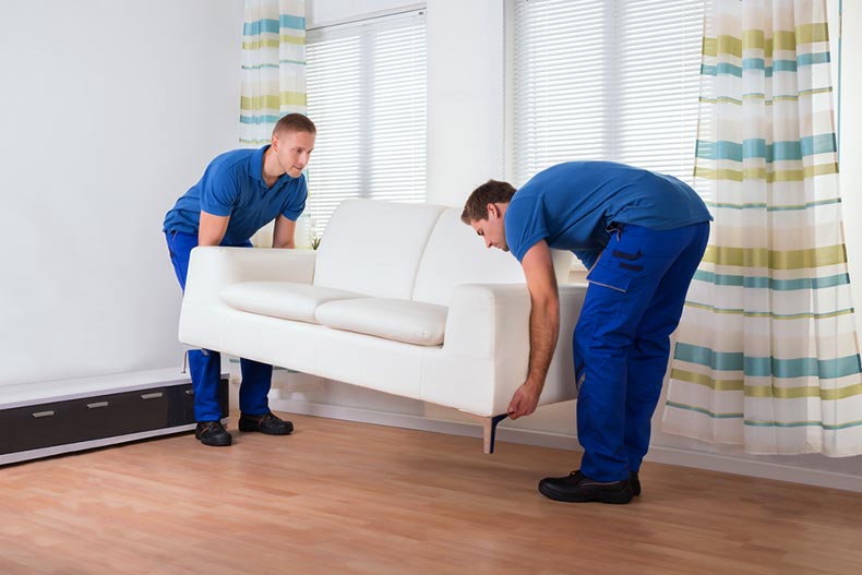 Two men from a professional staging service placing a white couch on a hardwood floor in a home for sale