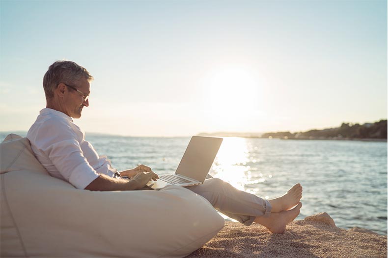 A senior man working on his laptop while sitting by the beach at sunset