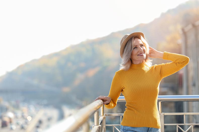 An older woman posing in the sunshine on a balcony with a city highway in the background