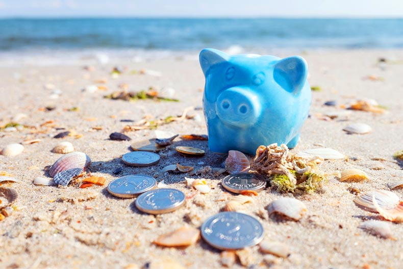 A blue piggy bank surrounded by coins on a tropical, sandy beach
