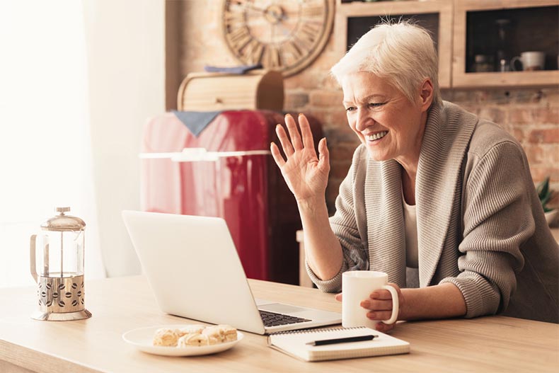 Elderly woman making a video call on a laptop in the kitchen and waving at the screen