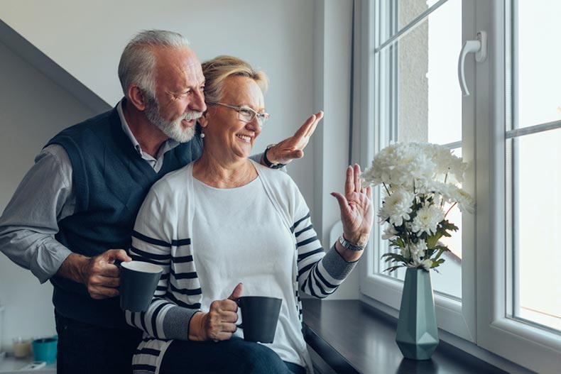 Elderly couple looking out the window while smiling and waving