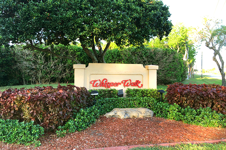 Community sign for Whisper Walk surrounded by red and green bushes and trees, as well as red mulch, located in Boca Raton, Florida