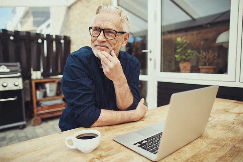 A senior man with a cup of coffee pausing to think while he works on his laptop