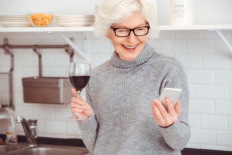 An old woman drinking wine while smiling and holding a smart phone